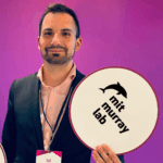 Headshot of Said Werner holding an MIT Murray Lab circular sign wearing a pink shirt and blue blazer in front of a purple background
